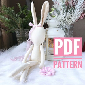 May include: A PDF pattern for sewing a white fabric bunny doll with long ears and a pink ribbon. The doll is sitting on a white fluffy surface with a pink flower.