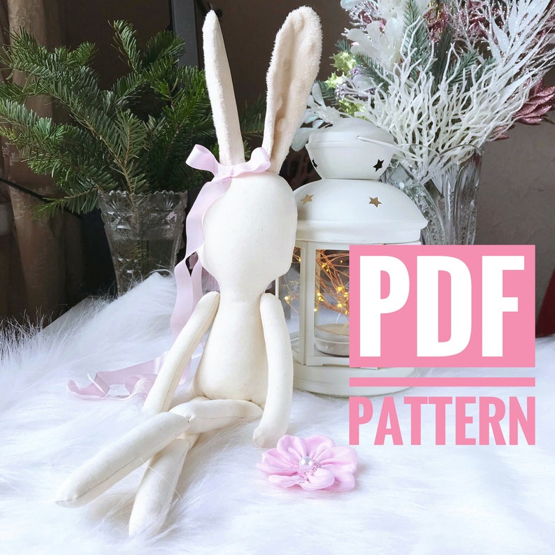 May include: A PDF pattern for sewing a white fabric bunny doll with long ears and a pink ribbon. The doll is sitting on a white fluffy surface with a pink flower.