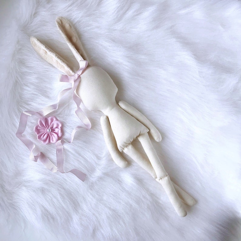 May include: A white fabric bunny doll with long ears and a pink ribbon around its head. The doll is lying on a white fluffy surface.