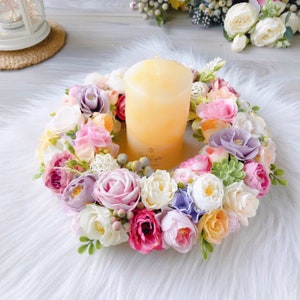 May include: A floral wreath with a variety of pastel-colored flowers, including pink, purple, white, and yellow, surrounds a yellow pillar candle. The wreath is on a white fluffy surface.