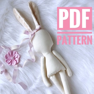 May include: A white fabric bunny doll with long ears and a pink ribbon around its neck. The doll is lying on a white fluffy surface. The text "PDF PATTERN" is written in pink letters on a white background.