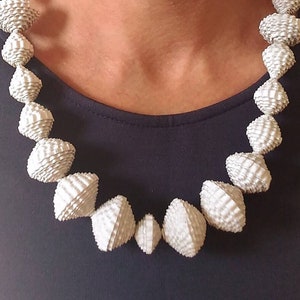 May include: An off-white necklace with geometric, textured beads. The beads are various sizes and shapes, creating a unique design. The necklace is worn against a dark blue top. The necklace's beads are made of a material that appears to be paper.