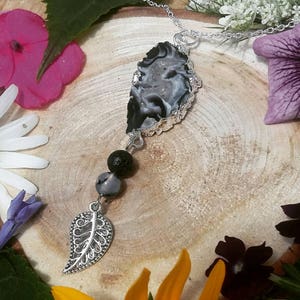 May include: A silver necklace featuring a large, irregularly shaped black and white stone pendant. Below the stone are three beads and a decorative silver leaf charm. The necklace is displayed on a wooden surface surrounded by flowers.