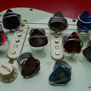 Può includere: Primo piano di una chitarra elettrica bianca con più plettri avvolti in filo d'argento. I plettri sono decorati con vari disegni e testi, tra cui "Wild About Music.Com" e "Texas".