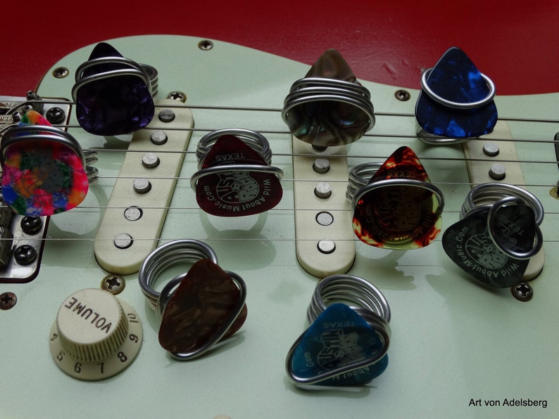 May include: A close-up of a white electric guitar with multiple guitar picks wrapped in silver wire. The picks are decorated with various designs and text, including "Wild About Music.Com" and "Texas".