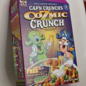 Puede incluir: Caja de cereales Cozmic Crunch de Cap'n Crunch. La caja presenta un extraterrestre verde y Cap'n Crunch vertiendo leche sobre los cereales. El texto indica que los cereales vuelven la leche verde y contienen Polvo Espacial Naranja. Peso neto 368g.
