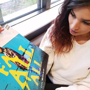 May include: A woman is holding a vinyl record with a blue and yellow cover. The record cover features a person wearing a plaid shirt and the text "Eazy-E" in yellow letters.