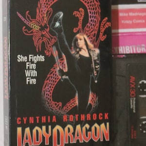 Puede incluir: Una cinta VHS de la película "Lady Dragon" protagonizada por Cynthia Rothrock. La portada presenta un dragón rojo y el texto "She Fights Fire With Fire".