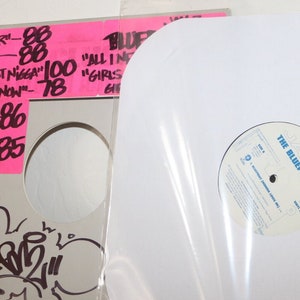 Puede incluir: Un disco de vinilo blanco en una funda de plástico transparente. La etiqueta del disco es blanca con texto azul que dice "The Blueprint".
