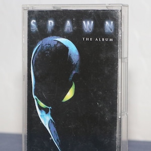 以下が含まれることがあります： 「Spawn: The Album」のヴィンテージカセットテープが表示されています。透明なカセットケースにはアルバムのアートワークが収められています。カバーには、青と黄色のアクセントが施されたSpawnの顔の暗い画像と、アルバムタイトルが描かれています。