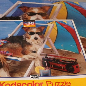 Puede incluir: Una caja de puzzle Kodacolor con un perro que lleva gafas de sol y una camisa a rayas rojas y azules, relajándose en una silla de playa con una radio y una sombrilla. La caja dice "Kodacolor Puzzle" y "550 piezas totalmente entrelazadas 45,7 cm x 61 cm Montado".