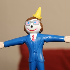 May include: A figurine of a cartoon character in a blue suit with a red tie, white gloves, and brown shoes. The character has a yellow party hat, glasses, and a smiling face. The figure is standing with arms outstretched.