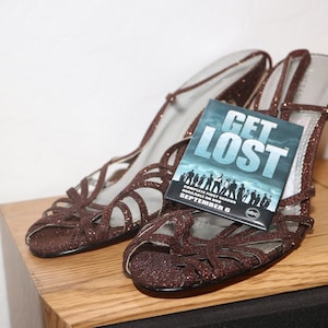 May include: A pair of brown sparkly heeled sandals with a "Get Lost" DVD box set on top. The DVD box set is blue with white text and features a group of people.