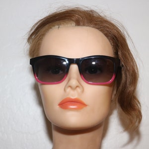 May include: Black sunglasses with pink accents on the frame. The sunglasses have a dark brown lens.