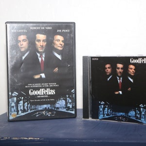 May include: A DVD case and a CD case for the movie "Goodfellas" are displayed. The DVD case features Ray Liotta, Robert De Niro, and Joe Pesci. The CD case has the same image. Both cases are on a blue surface.
