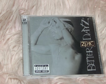 May include: A 2-CD album cover featuring a black and white photo of a man with a bandana on his head. The album title "Better Dayz" is printed in white on a black background. The artist's name "2PAC" is printed in white on a black background. The album cover has a parental advisory sticker.