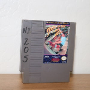 May include: A gray Nintendo Entertainment System (NES) game cartridge with the title "Skate or Die 2: The Search for Double Trouble" printed on the label. The label features a cartoon image of a skateboarder performing a trick.
