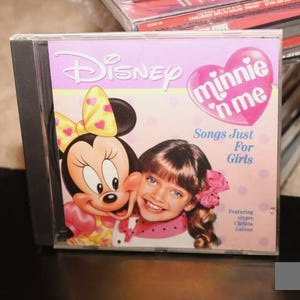 Puede incluir: Una caja de CD rosa y morada con una imagen de Minnie Mouse y una niña. El título del CD es "Disney Minnie 'n Me Songs Just For Girls" y presenta a la cantante Christina Larin.