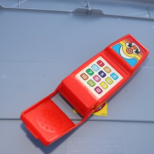 May include: A vintage red toy phone with a colorful keypad and a smiling face design. The phone has a handle and a dial pad with numbers 1-12. A classic toy for children.