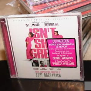 Może przedstawiać: Różowe i białe pudełko na płytę CD z obrazkiem Bette Midler i Nathana Lane'a na przodzie. Tytuł filmu to "Isn't She Great." Pudełko na płytę CD jest dla ścieżki dźwiękowej do filmu, która zawiera muzykę Burta Bacharacha. Tekst na pudełku głosi "Outrageous! Burt Bacharach is back with a sexy new soundtrack!"