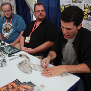 May include: Three people are sitting at a table signing autographs on books. The table is covered in a white tablecloth. There are several books on the table, including one with the title "Dark Eagle". A tie is lying on the table. The background is a wall with the words "Comic Con" printed on it.