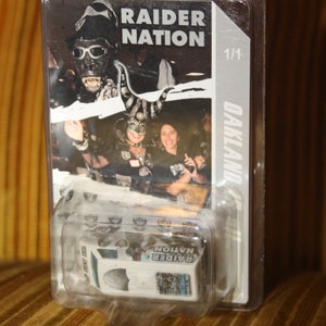Op de afbeelding: Een transparante plastic display met een zwart-witfoto van mensen die Raiders-kleding dragen. De foto heeft de tekst "Raider Nation" en "Oakland". De display bevat ook een klein wit doosje met de tekst "Raider Nation" en een afbeelding van een voetbalhelm.