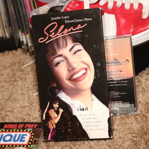 Könnte beinhalten: Eine VHS-Kassette des Films "Selena" mit Jennifer Lopez und Edward James Olmos. Das Cover zeigt ein lächelndes Porträt von Selena. Eine Musikkassette und andere Medien sind im Hintergrund zu sehen.