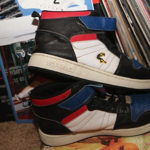 Puede incluir: Un par de zapatillas de deporte de caña alta negras, blancas, rojas y azules con la palabra "SHAG" en el lateral. Las zapatillas están encima de una revista.