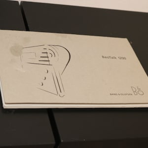 May include: A light gray booklet titled "BeoTalk 1200" with a stylized illustration of a telephone. The booklet has the Bang & Olufsen logo in the bottom right corner. The booklet is on a black surface.