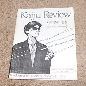 May include: A black and white magazine cover for Kaiju Review, Spring 1994, Volume One Number Five. The cover features a black and white illustration of a man in a suit and sunglasses holding a cigarette. The text "The Journal of Japanese Monster Culture" is at the bottom of the cover.