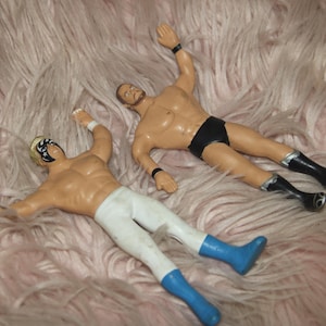 May include: Two wrestling action figures, one wearing a white and blue outfit and a black mask, the other wearing a black and white outfit. The figures are posed in a wrestling stance on a pink fluffy surface.