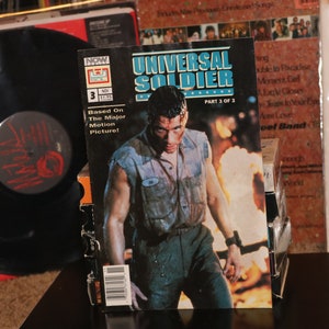 Op de afbeelding: Een comicboekomslag met een man in een vies, grijs, mouwloos shirt en blauwe jeans. De comic heet "Universal Soldier" en is deel 3 van 3. De comic is gebaseerd op de bioscoopfilm.