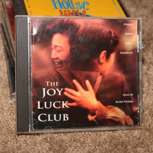 Peut inclure: Un boîtier de CD avec une image de fond rouge et orange de deux personnes qui s'embrassent. Le texte sur le devant indique "The Joy Luck Club" et "Bande originale du film".