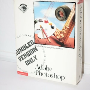 May include: A white box with a red and black logo for Adobe Photoshop. The box has a photo of a paint palette, a paintbrush, a camera lens, and a gold picture frame. The text on the box reads "Bundled Version Only" and "Adobe Photoshop".