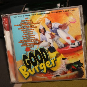 Pode incluir: Uma caixa de CD com um logotipo "Good Burger" amarelo e preto. A caixa apresenta uma foto de duas pessoas em um skate, com um hambúrguer no fundo. O texto na caixa diz "Música do filme original".