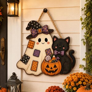 May include: A decorative wooden wall hanging featuring a cream-colored ghost wearing a polka-dot witch hat and a purple bow, standing next to a black cat with a purple bow, both positioned behind an orange jack-o'-lantern filled with candy.