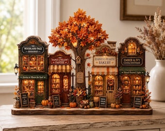May include: A miniature autumn village display featuring four shops: The Woodland Page Bookshop, Maple & Willow Pantry, Harvest Hearth Bakery, and Ember & Ivy Candle Boutique. A large central tree with orange leaves connects the buildings.