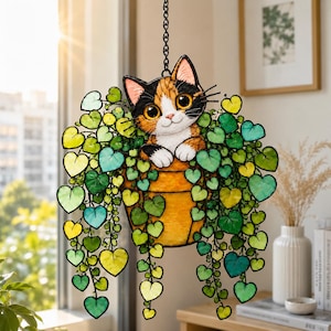 May include: A stained glass suncatcher featuring a calico cat with large eyes peeking out from a yellow pot. The cat is surrounded by cascading vines with numerous heart-shaped leaves in various shades of green and yellow, suspended by a black chain.