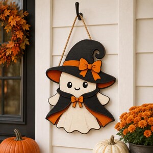 May include: A wooden wall hanging shaped like a ghost wearing a black witch hat with an orange bow and a black cape with an orange bow. The ghost is cream-colored with black eyes and a smile, suspended by a tan rope from a black metal hook.