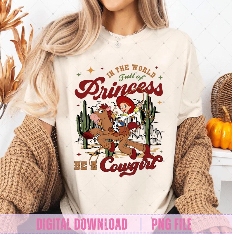 May include: Cream-colored t-shirt featuring a cartoon cowgirl riding a horse, with the text "In the world full of Princesses, Be a Cowgirl." The design includes cacti and stars in a Western theme.