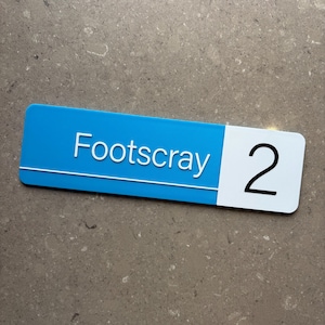 Op de afbeelding: Een rechthoekig bord met een blauw bovenstuk en een wit onderste gedeelte. Het woord "Footscray" staat in het wit en het nummer "2" in het zwart. Het bord is waarschijnlijk een huisnummer of adresmarkering.