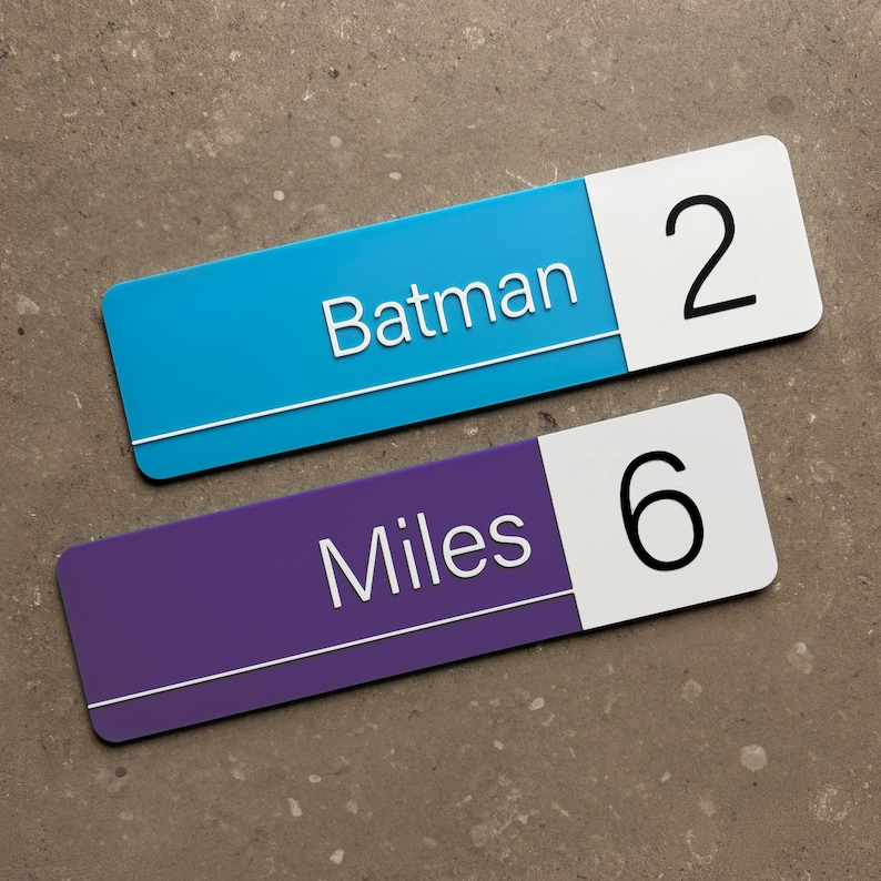 Op de afbeelding: Twee rechthoekige borden, &eacute;&eacute;n blauw en &eacute;&eacute;n paars, met witte tekst en cijfers. Het blauwe bord leest "Batman" en heeft het nummer "2". Het paarse bord leest "Miles" en heeft het nummer "6".