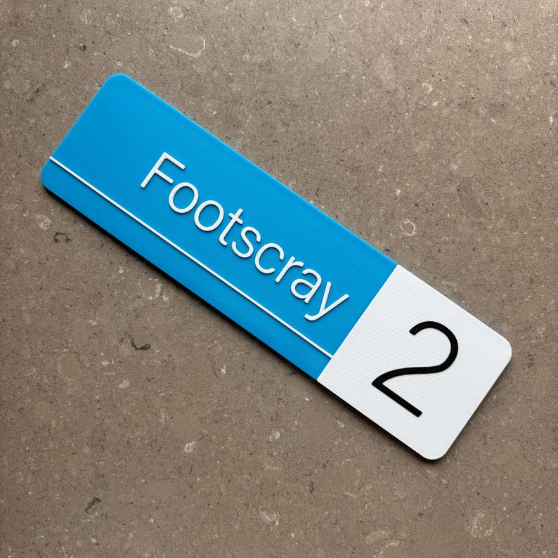 Op de afbeelding: Een rechthoekig bord met een blauw bovenstuk en een wit onderstuk. Het woord "Footscray" staat in het wit op het blauwe gedeelte en het nummer "2" staat in het zwart op het witte gedeelte.