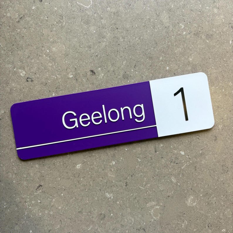 Op de afbeelding: Een rechthoekig bord met een paarse basis en een wit gedeelte. Het paarse gedeelte heeft het woord "Geelong" in het wit, en het witte gedeelte heeft het nummer "1" in het zwart. Het bord ligt op een grijs oppervlak.
