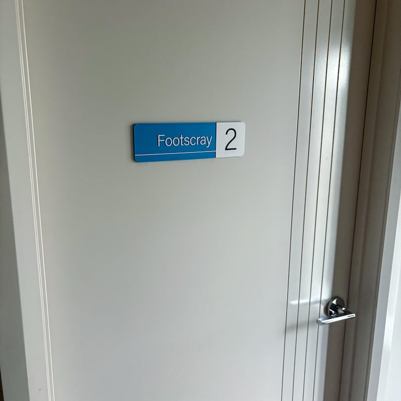 Op de afbeelding: Een rechthoekig bord met het woord "Footscray" in witte tekst op een blauwe achtergrond en het nummer "2" op een witte achtergrond. Het bord is op een witte deur gemonteerd.