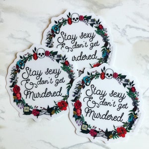 May include: Three round stickers with a floral wreath design. The wreath is made up of flowers, leaves, and other objects like a skull, a gun, and a bottle. The text on the stickers reads "Stay sexy & don't get murdered".