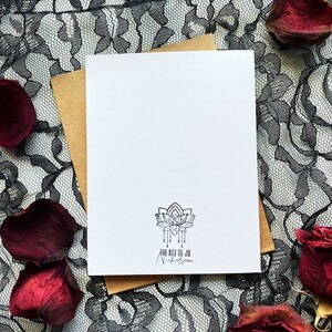May include: A blank white card with a brown envelope behind it. The card has a black and white logo with a stylized lotus flower and the text "The Art of Jo Nicholson". The card is on a black lace background with red rose petals.