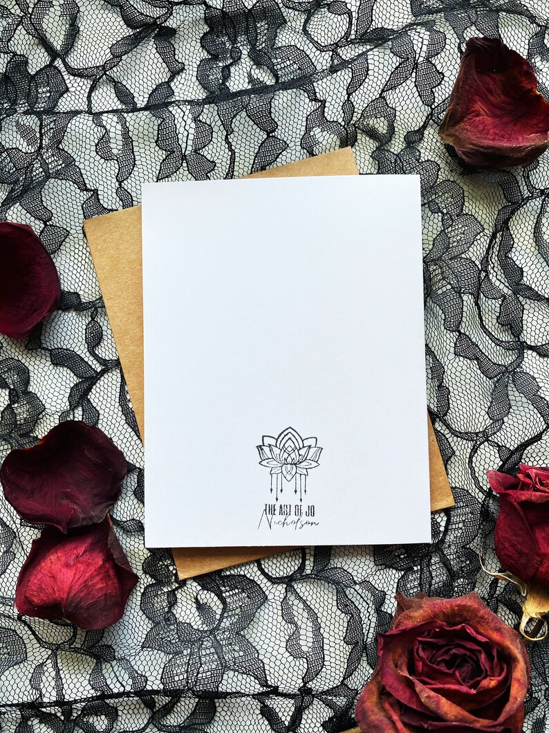 May include: A blank white card with a brown envelope behind it. The card has a black and white logo with a stylized lotus flower and the text "The Art of Jo Nicholson". The card is on a black lace background with red rose petals.