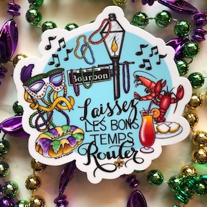 May include: A colorful sticker with a blue background and the text "Laissez Les Bons Temps Rouler" in black script. The sticker features a street lamp, a king cake, a crawfish, a hurricane cocktail, and a Mardi Gras mask.