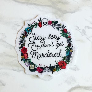 May include: A white sticker with a floral wreath design. The wreath includes a skull, a gun, a knife, a syringe, and a bottle of poison. The text inside the wreath reads "Stay sexy & don't get murdered".